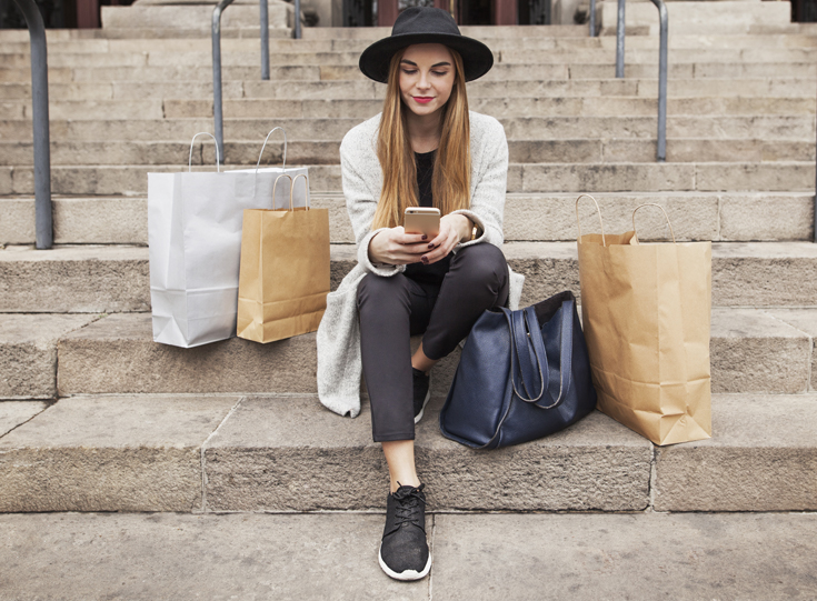Woman sitting on steps looking at her phone surrounded by shopping bags