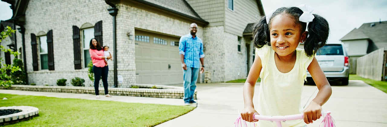 Little girl riding a scooter outside her home while her parents look on