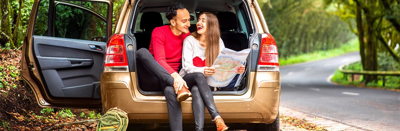 Young couple in sweaters sitting in the car trunk with paper map on the roadside in the forest. Young family traveling by car