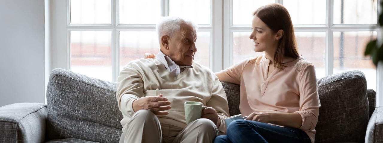 Smiling senior 80s man relaxing on sofa, talking to grown up daughter in living room. Happy different generations family enjoying drinking morning coffee with pleasant conversation together indoors.