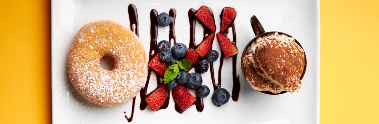 A mug of coffee and a powered sugar donut served with berries and a chocolate drizzle