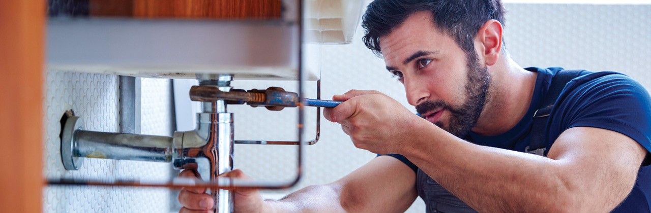 Male Plumber Using Wrench To Fix Leaking Sink In Home Bathroom