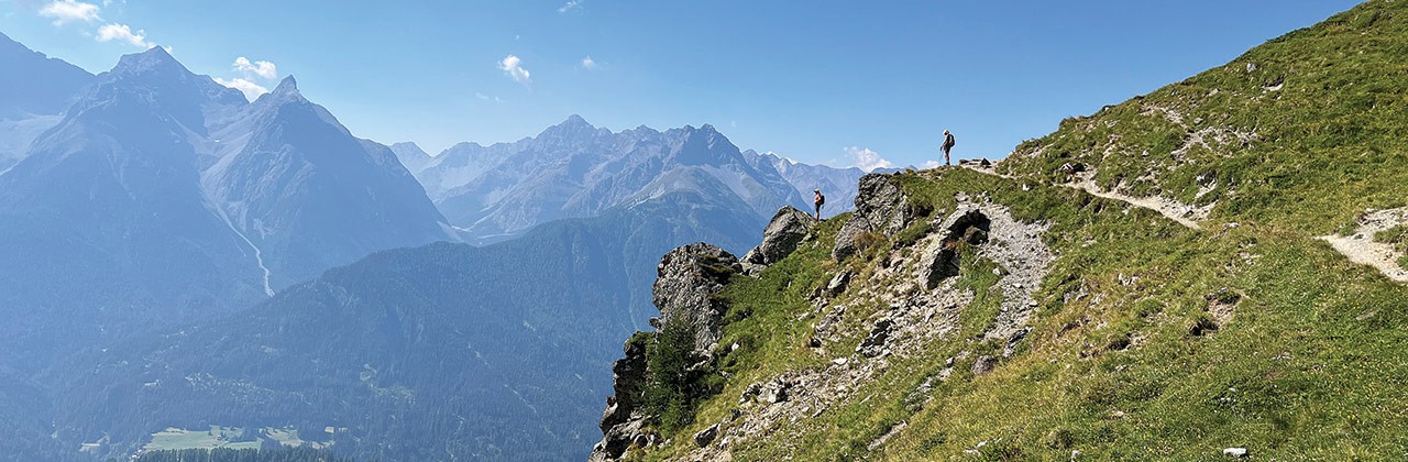 Two hikers stand on the side of a mountain, taking in sweeping views of the Engadin valley