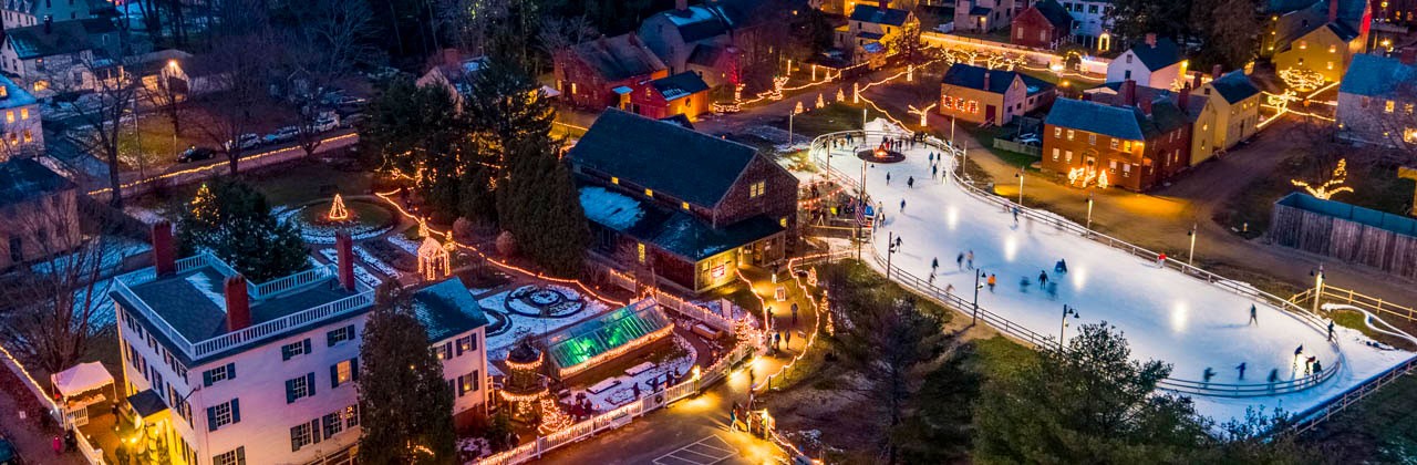 Aerial view of Labrie Family Skate at Puddle Dock Pond, the seasonal outdoor ice-skating rink, the grounds, and historic houses at Strawbery Banke Museum decorated for Candlelight Stroll.