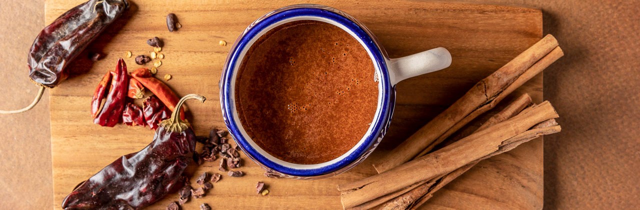 A mug of hot chocolate flanked by roasted chiles and cinnamon sticks