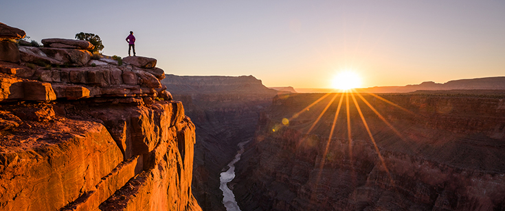 A hiker stands at the endge of a cliff as the sun rises in Grand Canyon National Park, Arizona