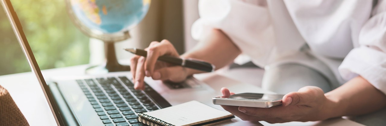 A woman doing travel planning with a laptop, pen, notepad, and phone