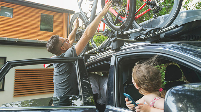 A father loads bicycles on top of an SUV while his daughter watches
