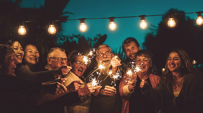 A group of people hold sparklers under a string of lights in the evening