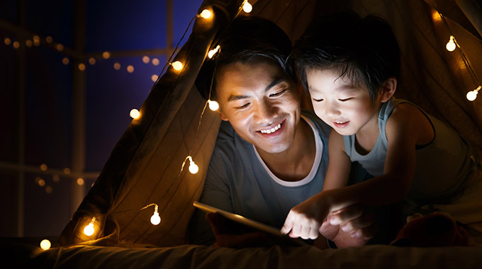 A father and son use a tablet while lying in a tent indoors
