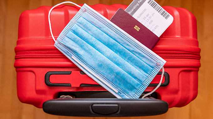 A surgical face mask on top of a passport with a boarding pass and a red suitcase