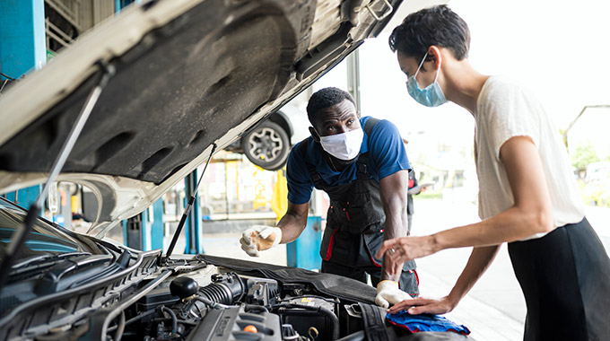 A mechanic or service rep talks to a female customer, both wearing masks, under the hood of a car