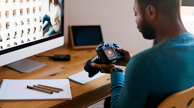 A man holds a DSLR camera while sitting at a computer