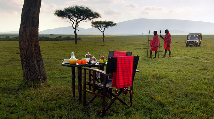 Two chairs at a small table hosting a "bush breakfast" on an Kenyan safari, with two Maasai warriors in the background