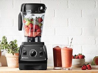 A Vitamix blender with fruit inside, next to 2 smoothies, on a kitchen counter