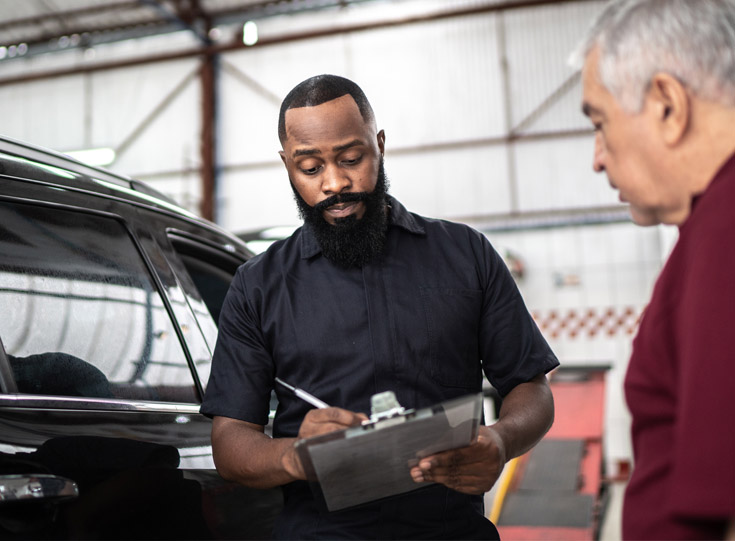 Mechanic reviewing paperwork while standing between car and customer Mechanic reviewing paperwork while standing between car and customer