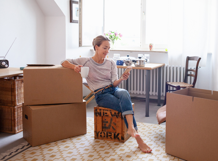 A woman relaxes with coffee and browses her cellphone while sitting among packing boxes A woman relaxes with coffee and browses her cellphone while sitting among packing boxes