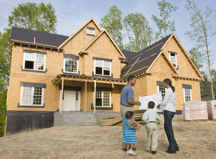 Couple with two children standing in front of a home construction looking at blueprints Couple with two children standing in front of a home construction looking at blueprints