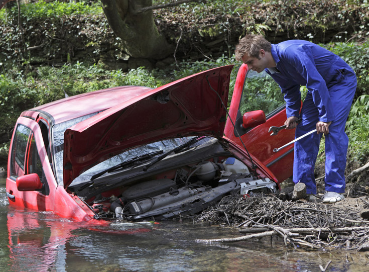 Man in coveralls with tools looking at a car submerged in water Man in coveralls with tools looking at a car submerged in water