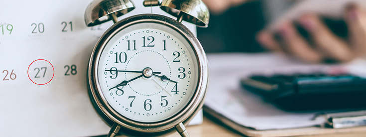 An alarm clock next to a calendar with a 27 circled in red and a blurred out desk on the right with a calculator and a hand. An alarm clock next to a calendar with a 27 circled in red and a blurred out desk on the right with a calculator and a hand.