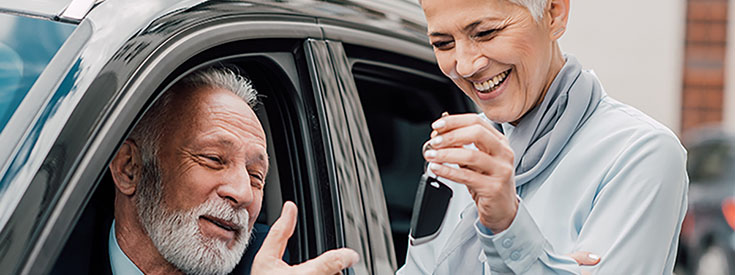 Senior woman hands senior man key to car he's sitting in. Senior woman hands senior man key to car he's sitting in.