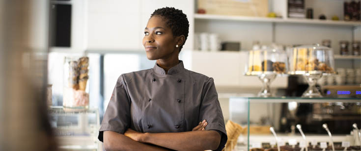 Chef dressed in gray stands in front of a case of baked goods Chef dressed in gray stands in front of a case of baked goods