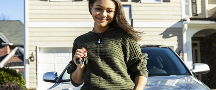 A young driver standing in front of her car smiles while holding up the keys A young driver standing in front of her car smiles while holding up the keys