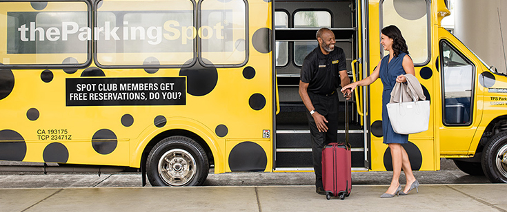 An employee from The Parking Spot assisting a woman with her suitcase in front of a shuttle bus An employee from The Parking Spot assisting a woman with her suitcase in front of a shuttle bus