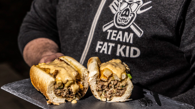 Someone wearing a "Team Fat Kid" hoodie holds up a tray with a sliced Best Philly