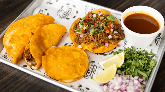 A tray with birria tacos, birria quesadillas, consomme, and lime, cilantro, and onions for garnish. 