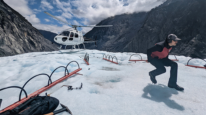 Man walking on Fox Glacier, a helicopter perched on the edge of the formation.