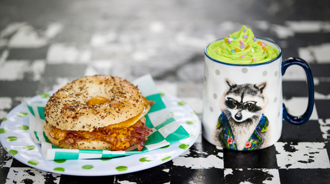 Bagel sandwich with bacon and egg beside a mug of coffee topped with whipped cream and sprinkles.