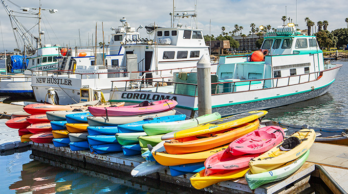 Kayaks piled on a dock in Oxnard, with fishing boats in the background.