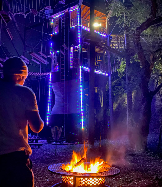A visitor staying warm by a fire near a treetop structure at Coral Crater Adventure Park.