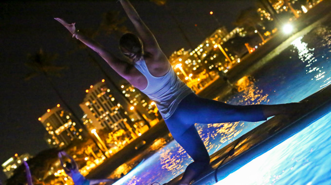 A woman reaching for the sky during a nighttime paddleboard yoga class.