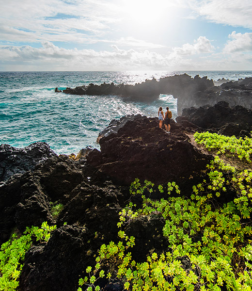 Hikers at Wai‘ānapanapa State Park take in a view of the dramatic volcanic coastline and a natural stone arch. | Photo by Tommy Lundberg/Hawaii Tourism Authority