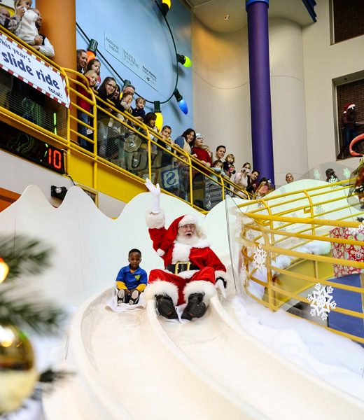 A young boy and Santa gliding side by side down the Yule Slide at The Children's Museum of Indianapolis