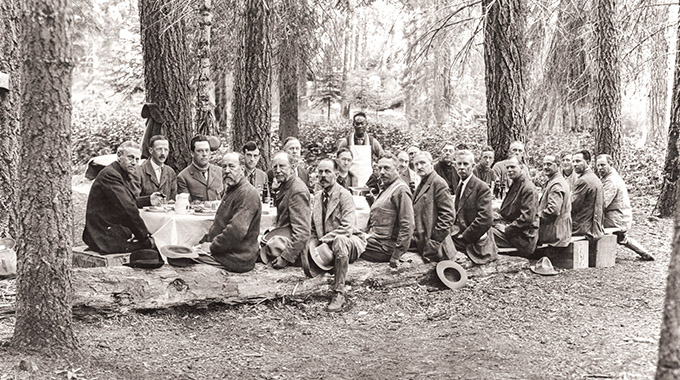 Tie Sing (in apron) presides over a meal he prepared during the 1915 Mather Mountain Party Expedition. Seated at the far left is Stephen T. Mather, who became the first director of the National Park Service. | Photo by Dr. Gilbert H. Grosvenor/National Geographic Creative