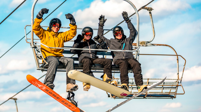Three snowboarders raising their arms while riding a ski lift at Canaan Valley Resort State Park