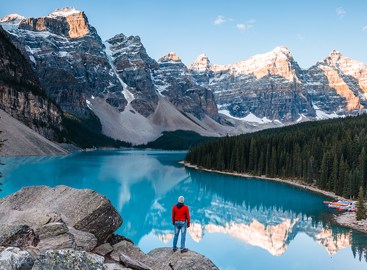 Man in red jacket standing at cliffside over crystal-clear water reflecting mountains in the background Man in red jacket standing at cliffside over crystal-clear water reflecting mountains in the background
