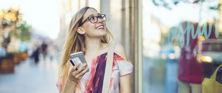 Woman in glasses walking by a shop window with cellphone and card in hand