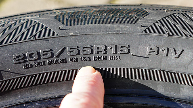 A finger points to the tire size and speed/load ratings on the sidewall of a car tire