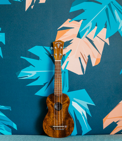 A ukulele resting against a wall with monstera leaf wallpaper